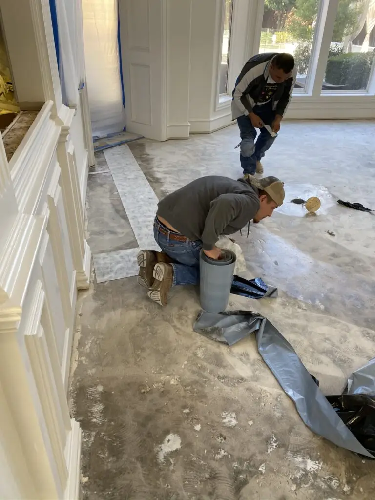 Two workers engaged in the process of removing ceramic tile from a floor, with one kneeling and the other standing in the background.