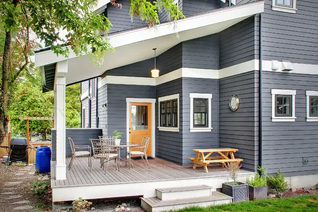 Exterior of a modern gray house with white trim and a prominent wooden deck featuring outdoor furniture, a covered porch, and a light orange front door. Green foliage is visible in the background.